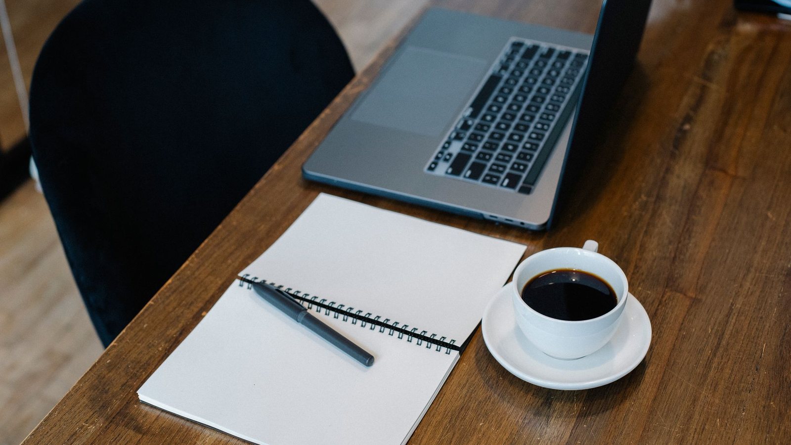 An open laptop, a notebook, a pen and a cup of coffee on a wooden desk, seen from above.