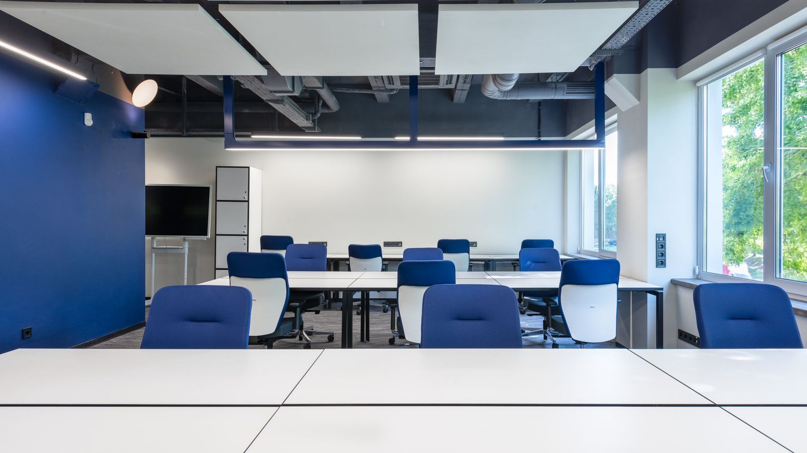 Rows of white desks with blue chairs in a bright, spacious office with a large window and a monitor.