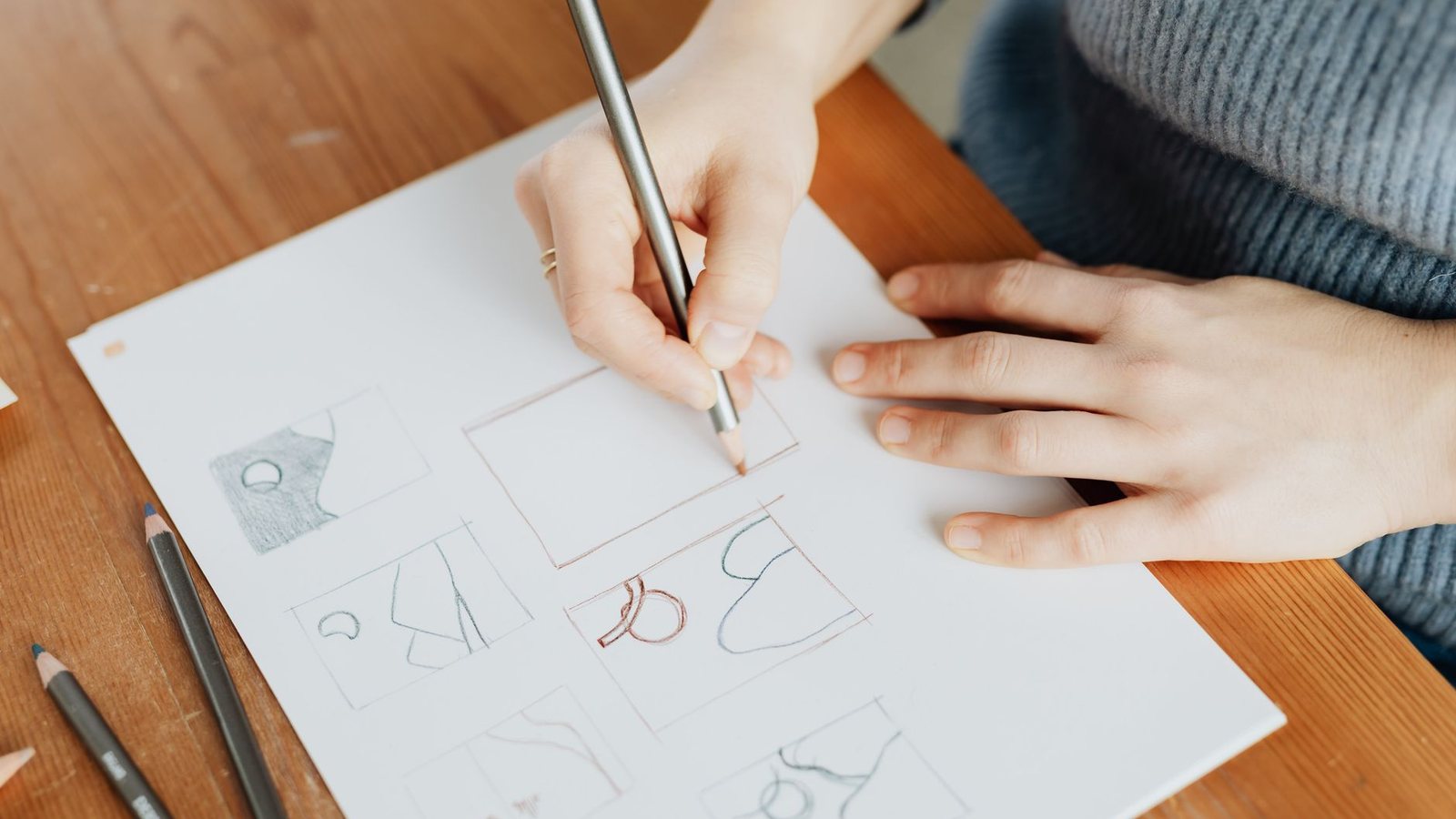 A designer at a wooden table drawing a mark in pencil on a sheet of paper, seen from above.